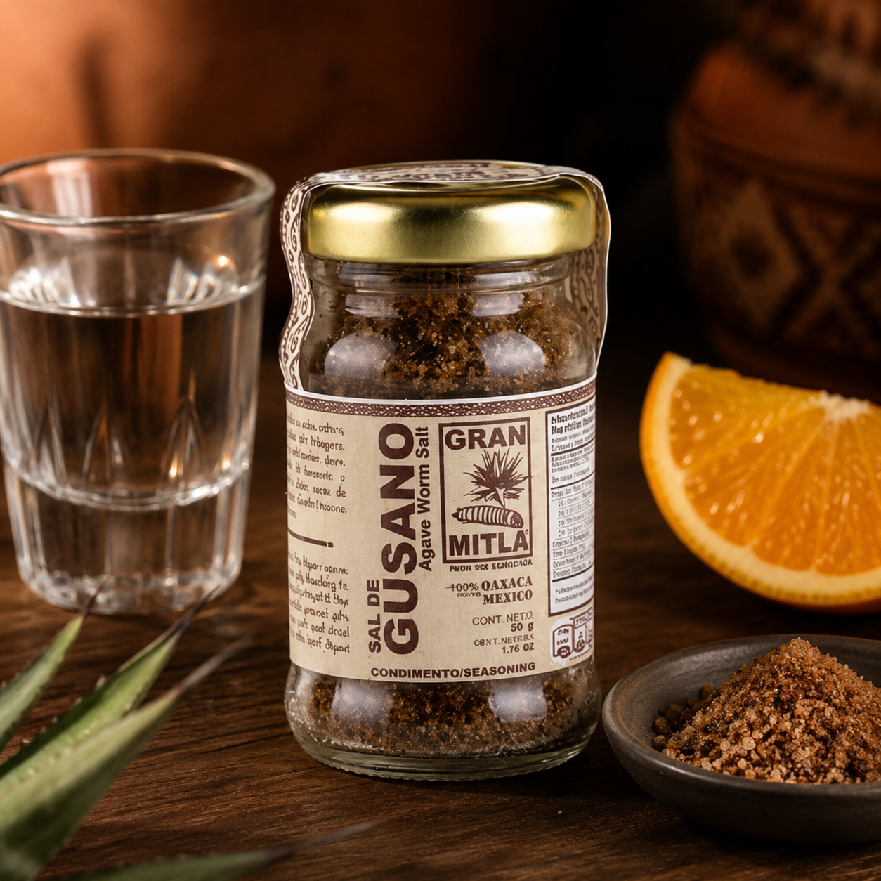 A jar of Gusano Agave Worm Seed by Mitla, a Mexican organic agave syrup producer, displayed on a rustic wooden table with a glass of water, a slice of orange, and a small bowl of the brown worm seed seasoning.
