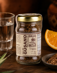 A jar of Gusano Agave Worm Seed by Mitla, a Mexican organic agave syrup producer, displayed on a rustic wooden table with a glass of water, a slice of orange, and a small bowl of the brown worm seed seasoning.