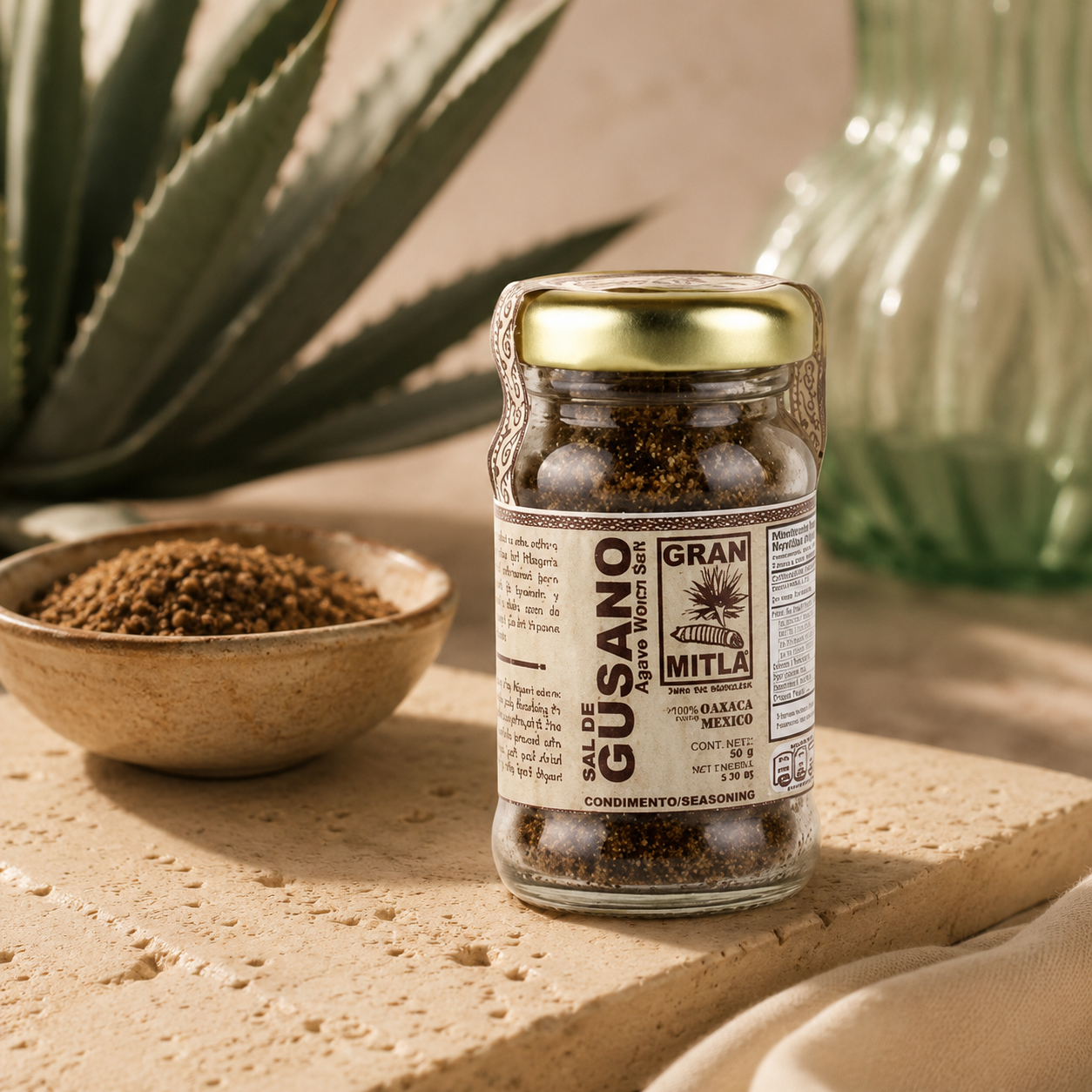 A glass jar of Gran Mitla Gusano Alawa Mole Salt with a gold lid and brown label, set on a stone surface next to a small bowl of the ground salt, with an aloe vera plant and a green glass bottle in the background.