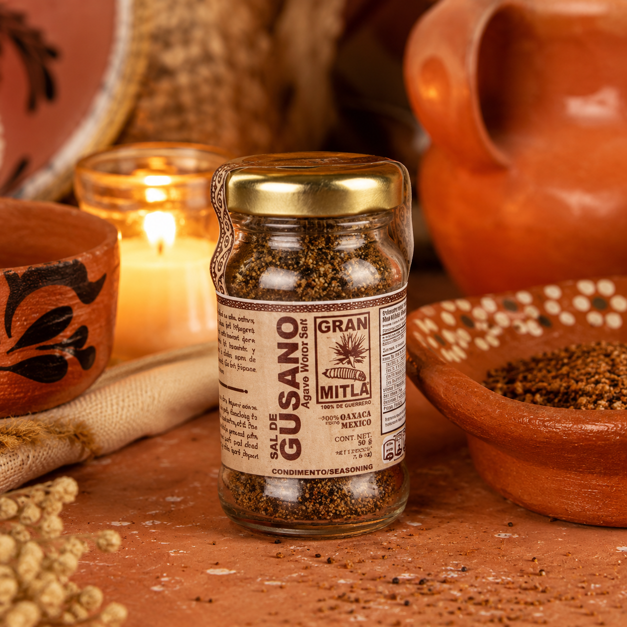 A glass jar of Gran Mitla Sal de Gusano seasoning, featuring a gold lid and a brown label, set on a rustic terracotta surface with a lit candle, bowls, and dried ingredients in the background.