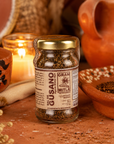 A glass jar of Gran Mitla Sal de Gusano seasoning, featuring a gold lid and a brown label, set on a rustic terracotta surface with a lit candle, bowls, and dried ingredients in the background.