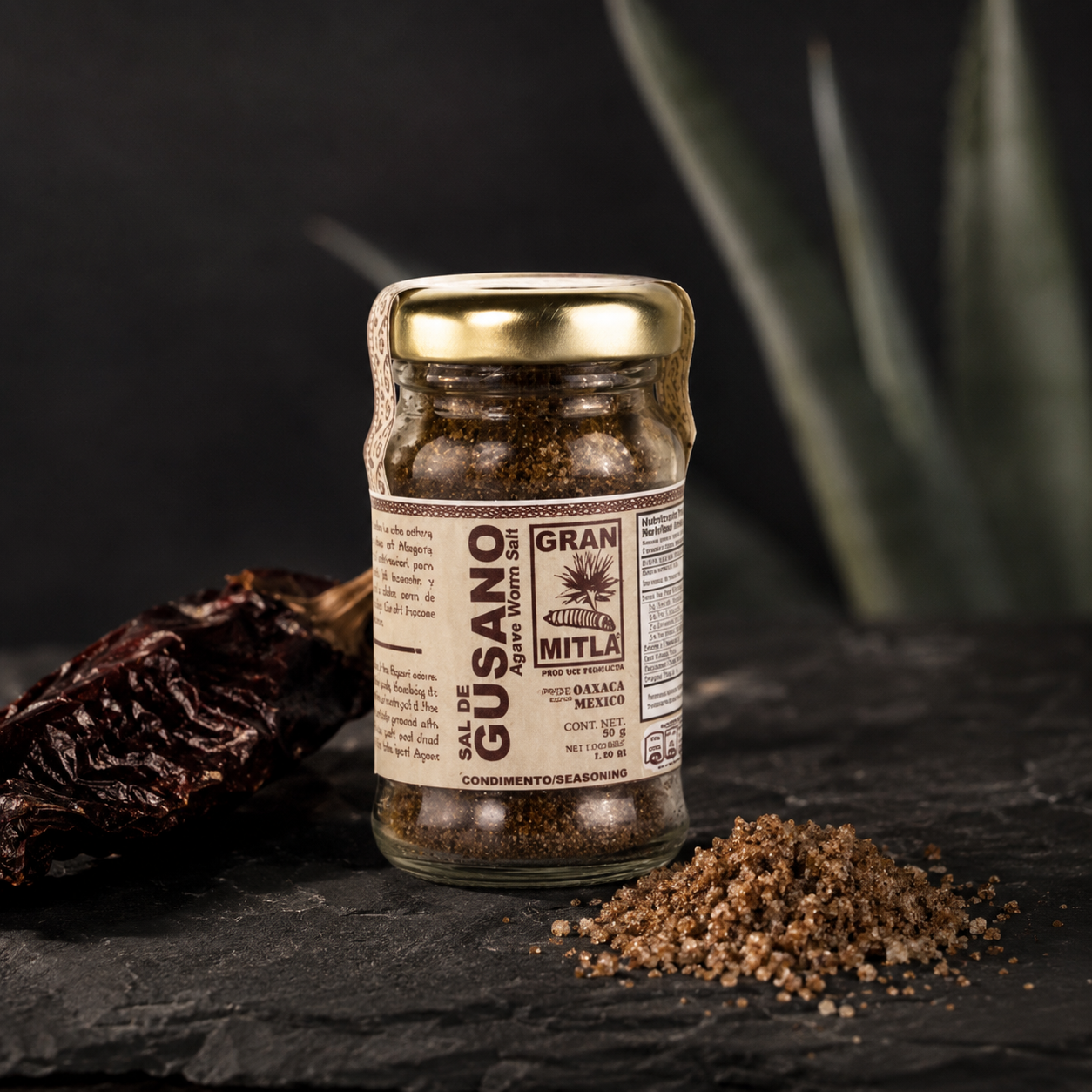 A glass jar of Gran Mitla Guasano seasoning blend with a gold lid, set on a dark slate with dried chiles and seasoning powder. The background features blurred green aloe leaves.