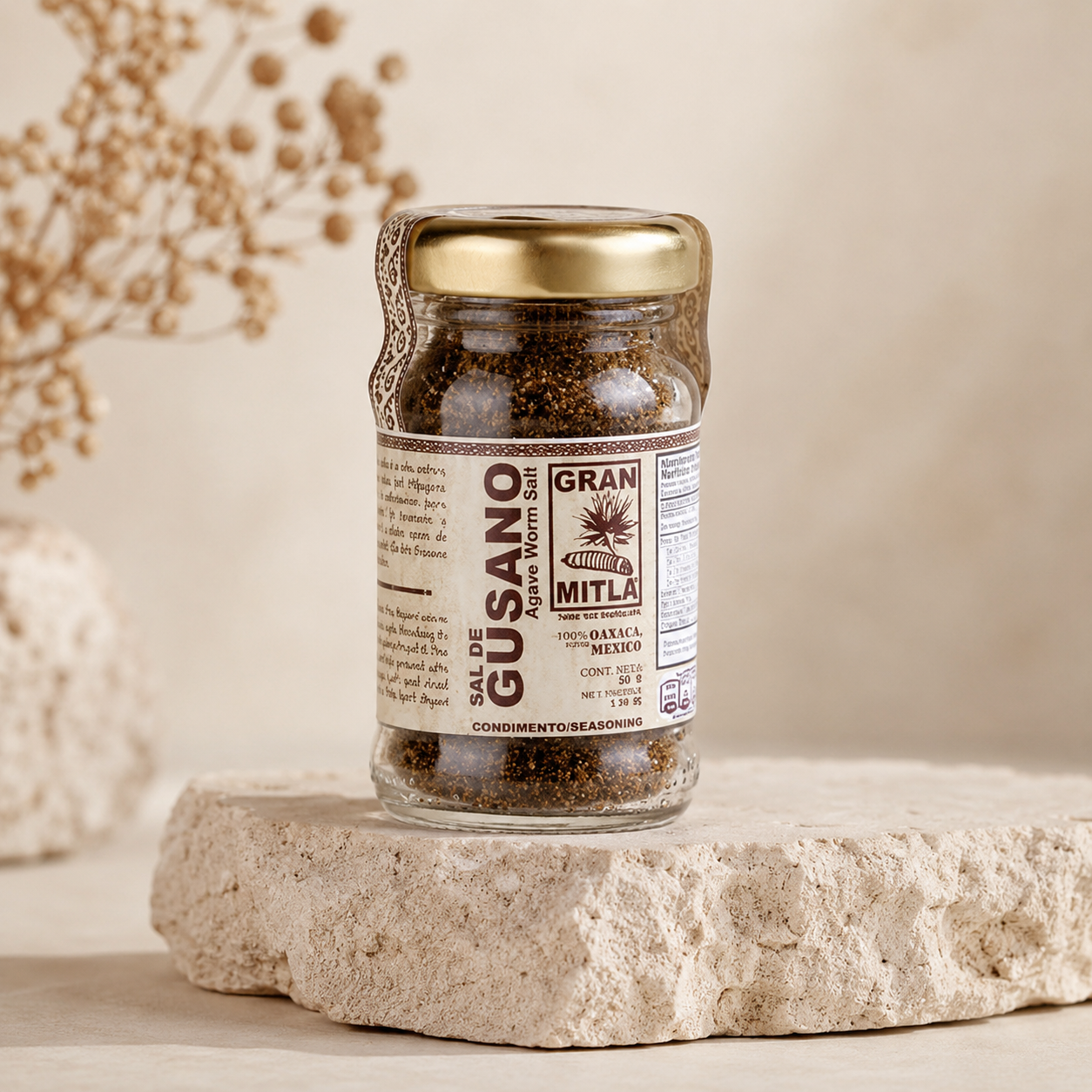 A glass jar of Gran Mitla Sal de Gusano (gourmet sea salt) with a gold lid and brown label, displayed on a textured stone pedestal against a warm beige background.