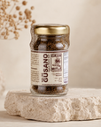 A glass jar of Gran Mitla Sal de Gusano (gourmet sea salt) with a gold lid and brown label, displayed on a textured stone pedestal against a warm beige background.