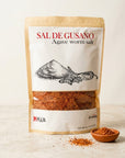 A stand-up pouch of Mexa Sal de Gusano (Agave Worm Salt) on a light beige background, with a small wooden bowl and scattered seasoning in front.
